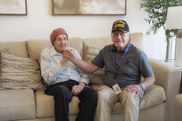 A man wearing a Vietnam Veteran hat holds hands with WWII Vet Ned Kent. They are sitting on a couch.