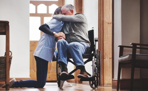 A caregiver and a person in a wheelchair are hugging. Their faces are obscured by their arms.