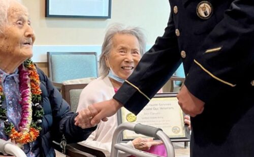 A young veteran is standing, holding the hand of a seated Veteran. The patient is smiling at him.