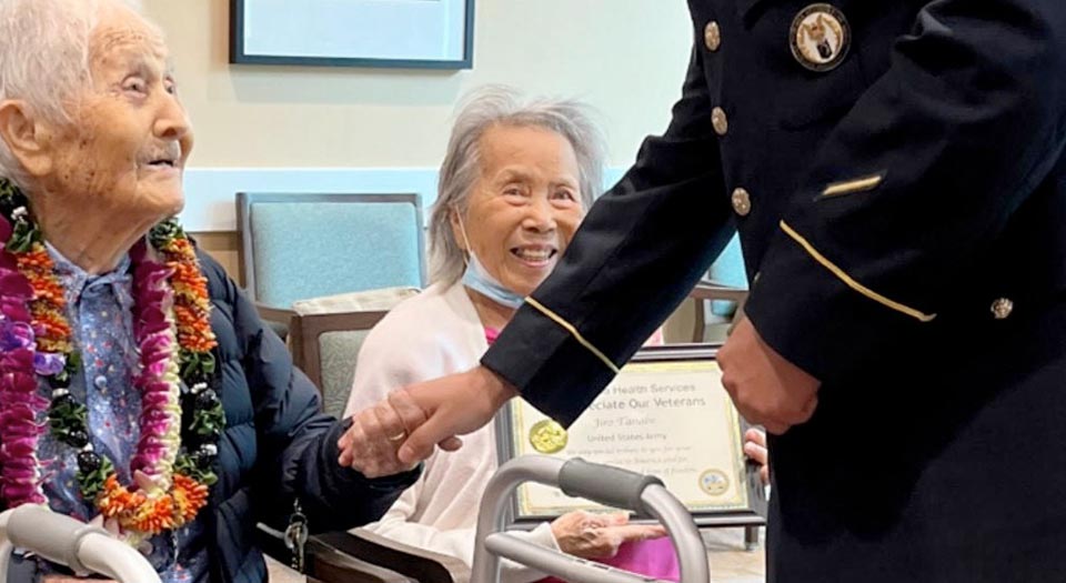 A young veteran is standing, holding the hand of a seated patient. The patient is smiling at him.